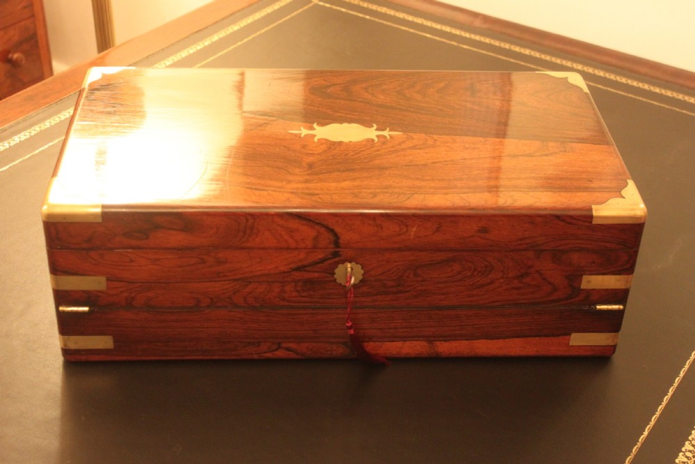 A rectangular dark rosewood writing slope box with brass corner mounts, brass cartouche inlay, and a central brass keyhole with a tassel.