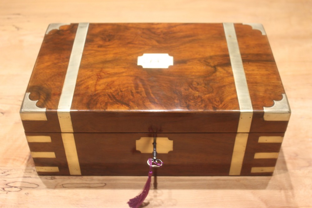 A closed Victorian figured walnut writing slope featuring protective brass straps, corner mounts, an inlaid central cartouche, and a key inserted into the lock.