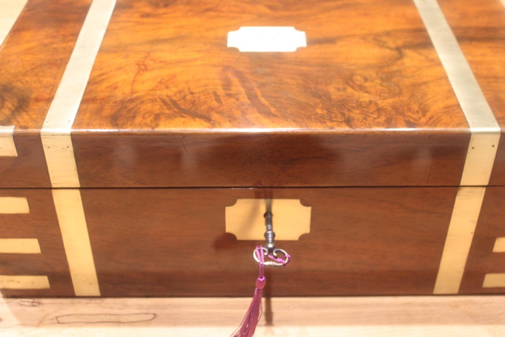 A closed Victorian figured walnut writing slope featuring protective brass straps, corner mounts, an inlaid central cartouche, and a key inserted into the lock. View II