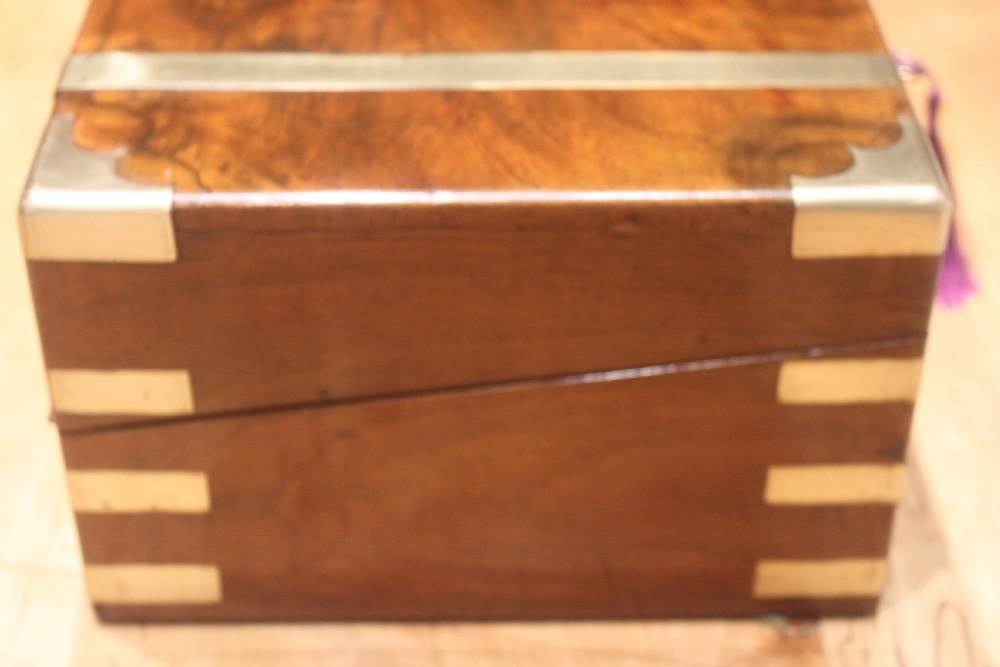 A closed Victorian figured walnut writing slope featuring protective brass straps, corner mounts, an inlaid central cartouche, and a key inserted into the lock. View III