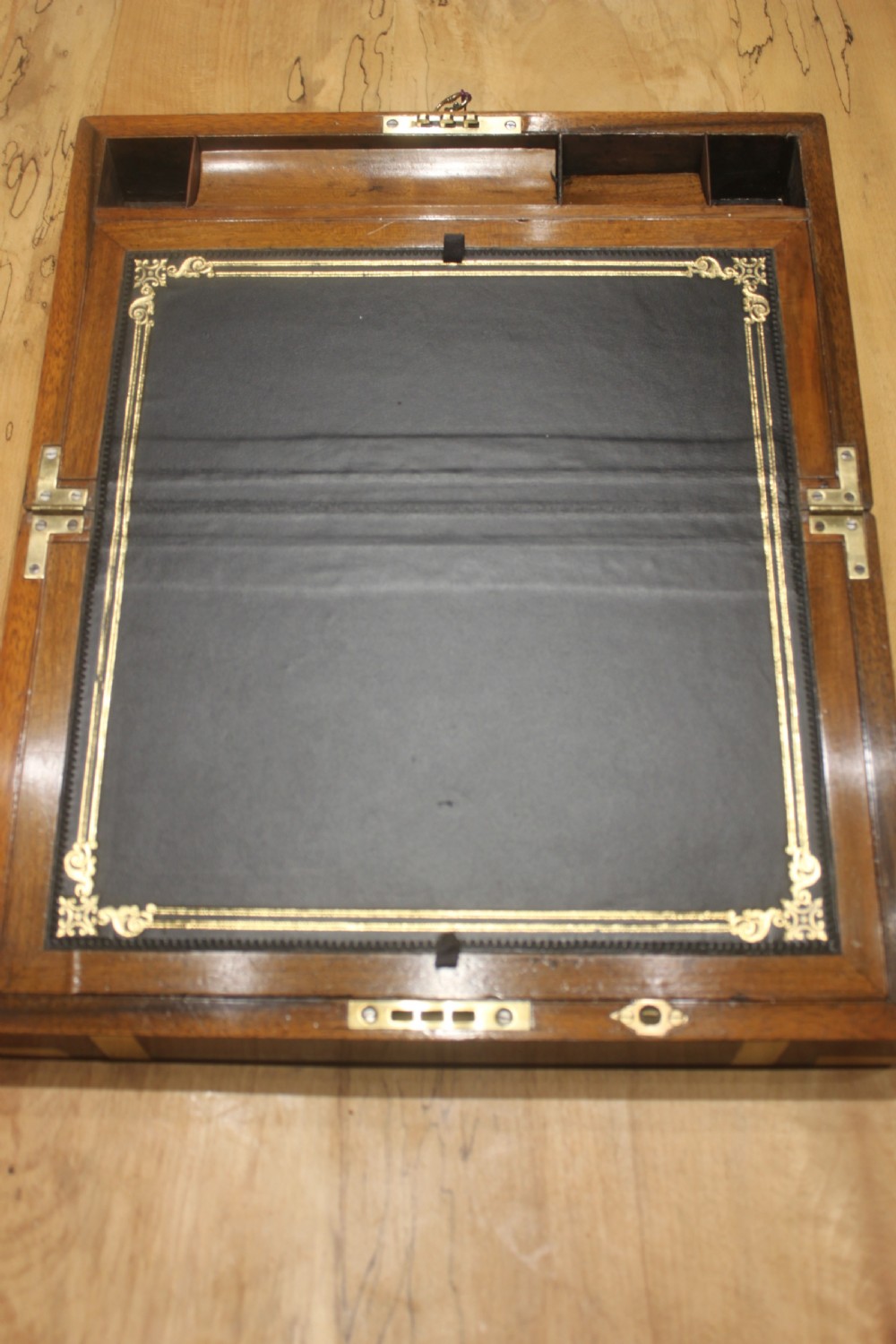 A closed Victorian figured walnut writing slope featuring protective brass straps, corner mounts, an inlaid central cartouche, and a key inserted into the lock. View VI