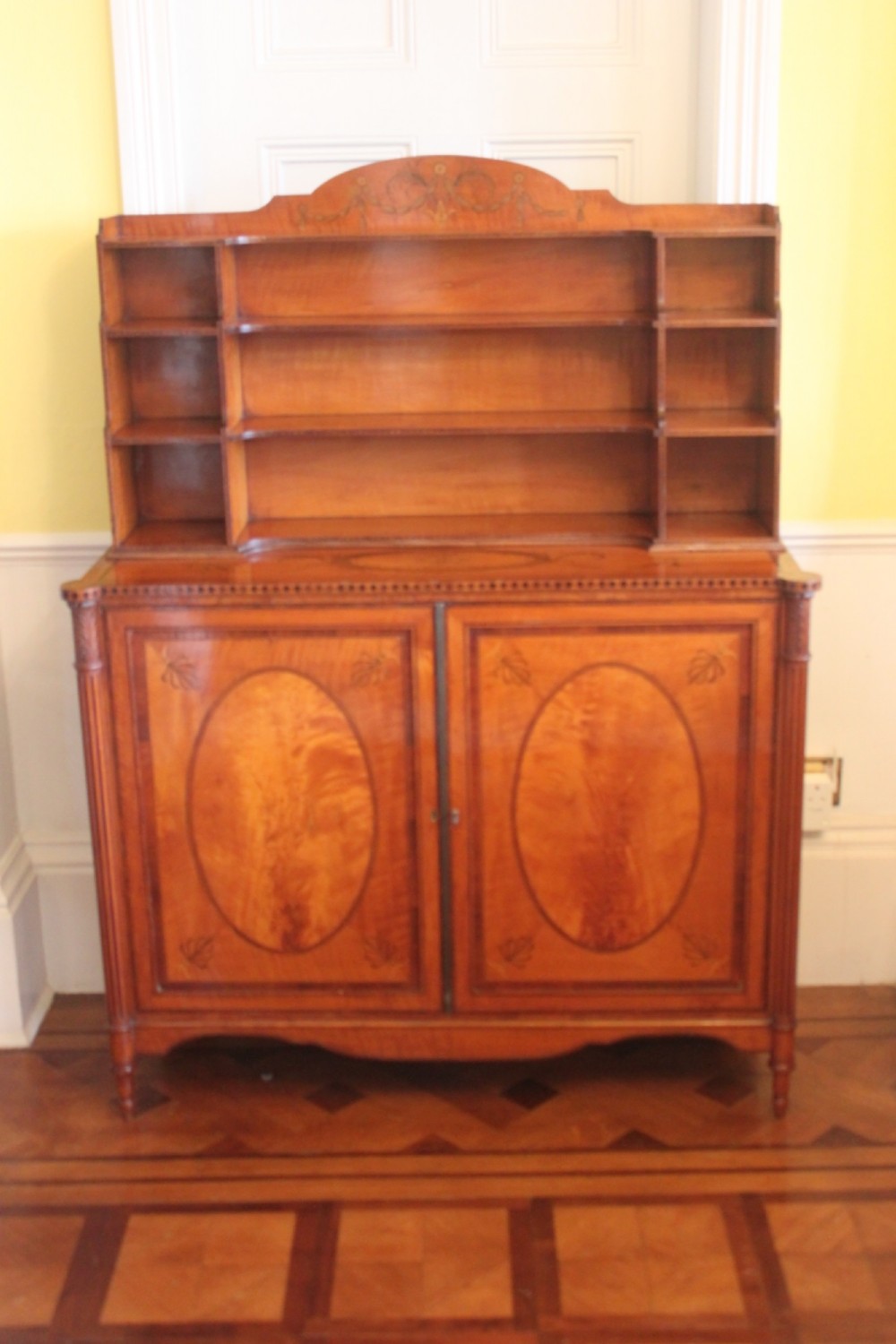 An antique Sheraton style satinwood cabinet with an open tiered shelving top unit and a lower two-door cabinet featuring oval inlaid marquetry panels.