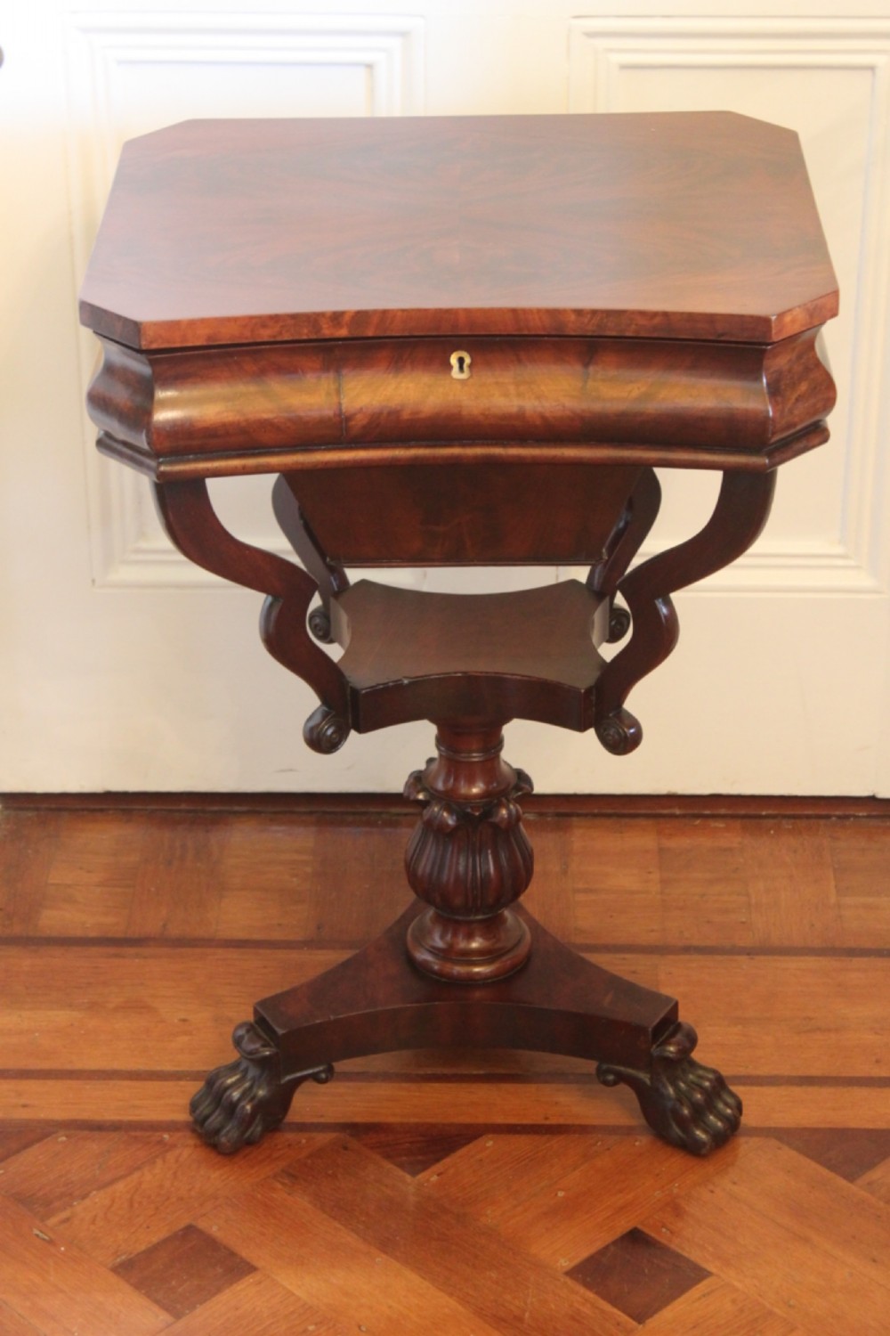 An early Victorian flame mahogany work table featuring a hinged top, carved central pedestal, triform base, and carved lion paw feet.