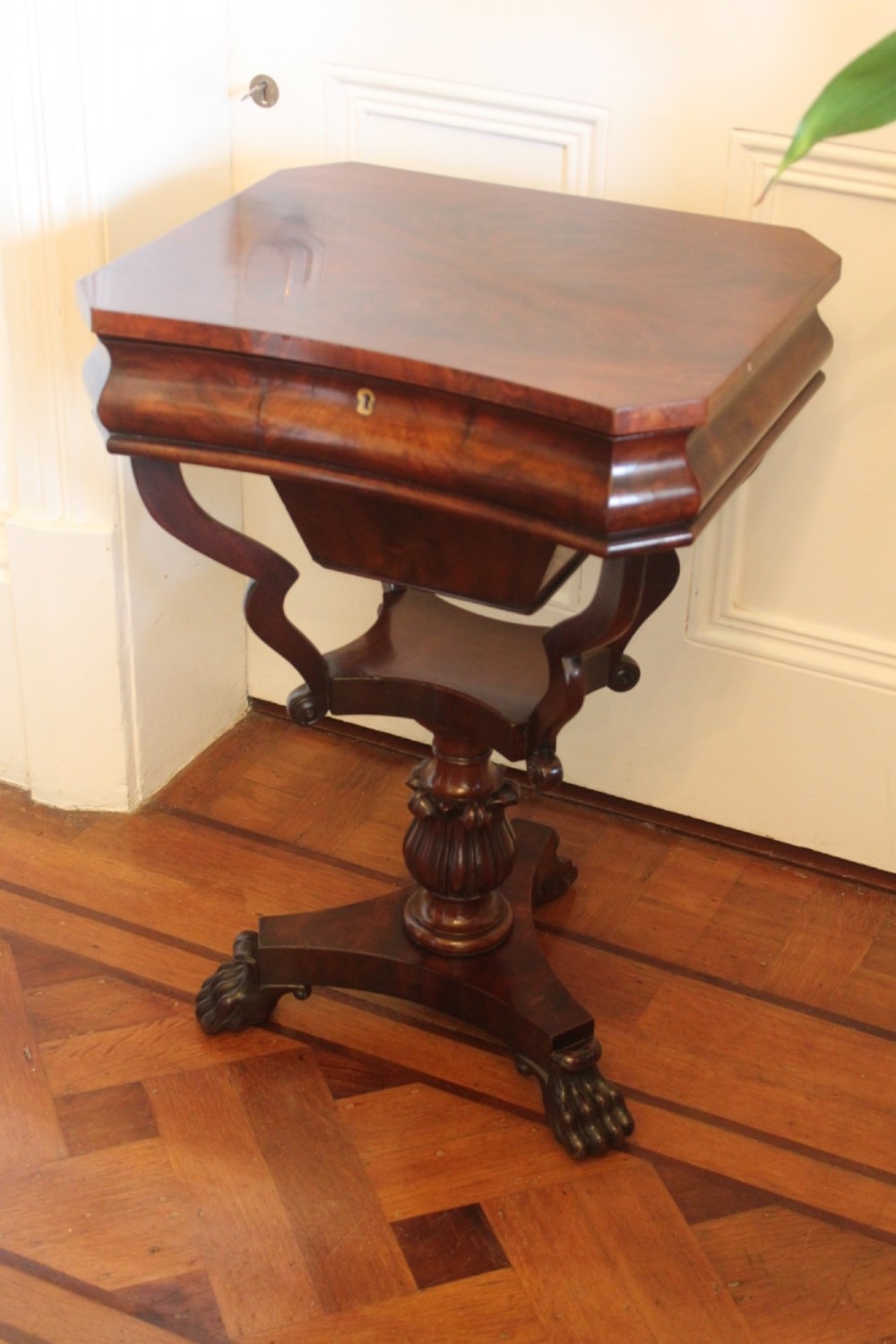 An early Victorian flame mahogany work table featuring a hinged top, carved central pedestal, triform base, and carved lion paw feet. View II
