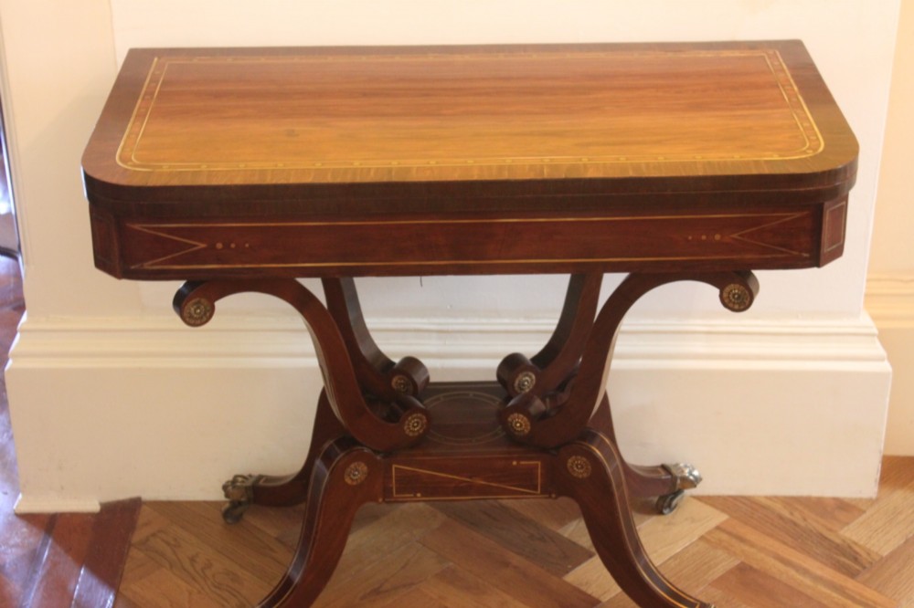 A Regency mahogany fold-over card table featuring geometric brass inlay, scroll supports, and brass paw feet on castors.