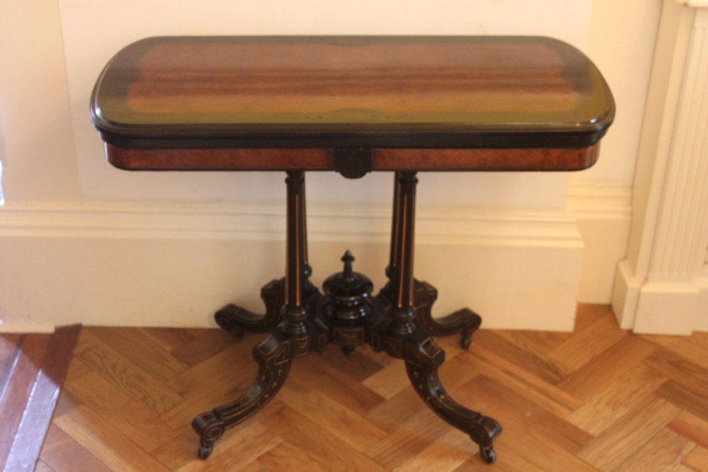 A Victorian foldover card table featuring a burr walnut top, ebonised border, double twin-column pedestal, and carved legs with castors.