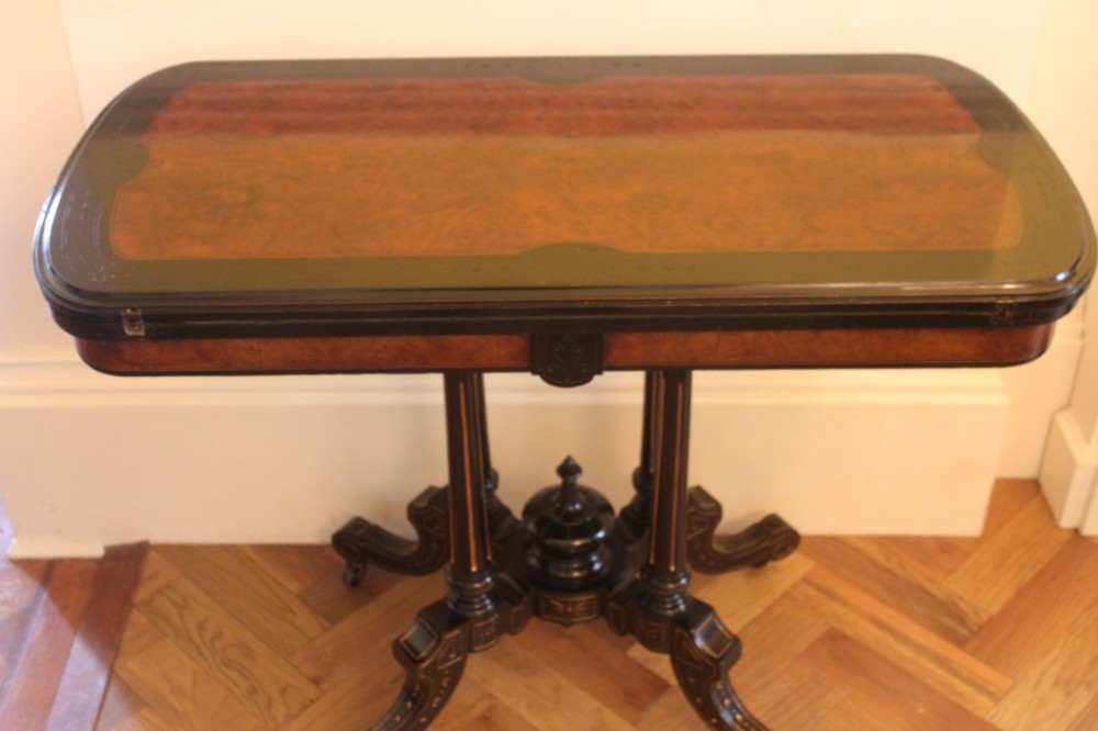 A Victorian foldover card table featuring a burr walnut top, ebonised border, double twin-column pedestal, and carved legs with castors. View XI