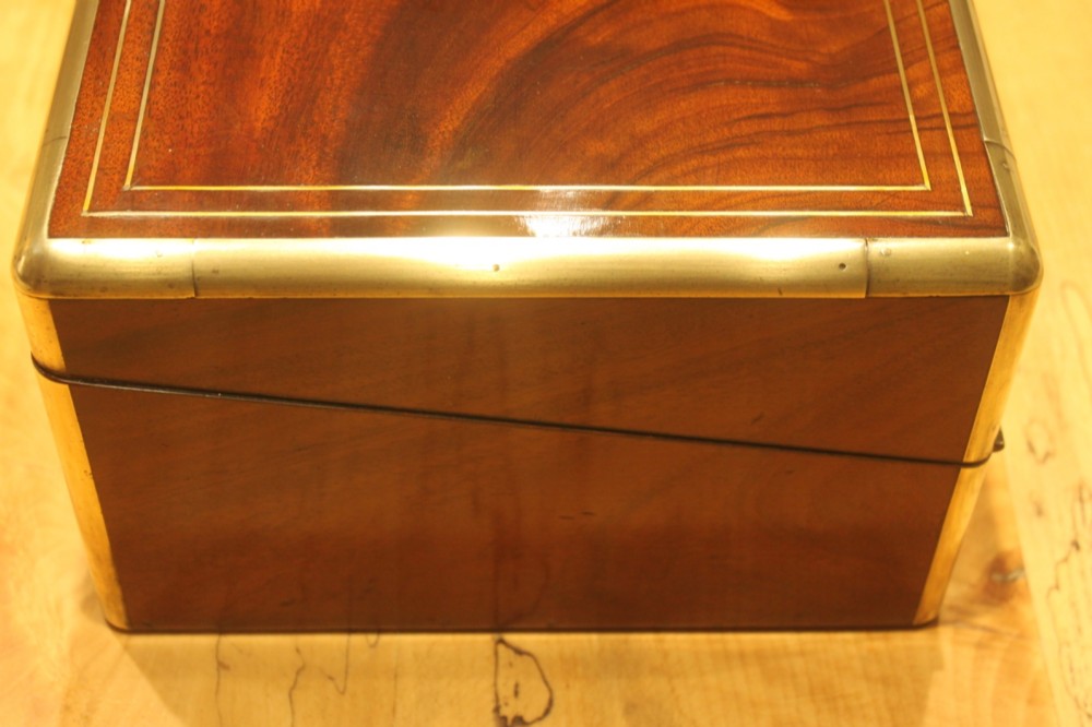 A rectangular curl grain mahogany writing slope box featuring fine brass stringing, brass bound corners, a central brass cartouche, and a keyhole escutcheon. View V