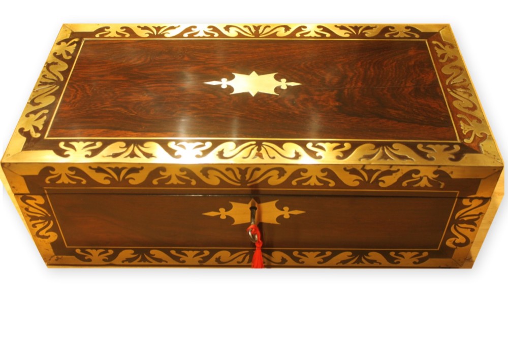 A closed rectangular dark rosewood writing slope box featuring detailed brass foliate inlay along the borders and a brass key inserted into the front lock.