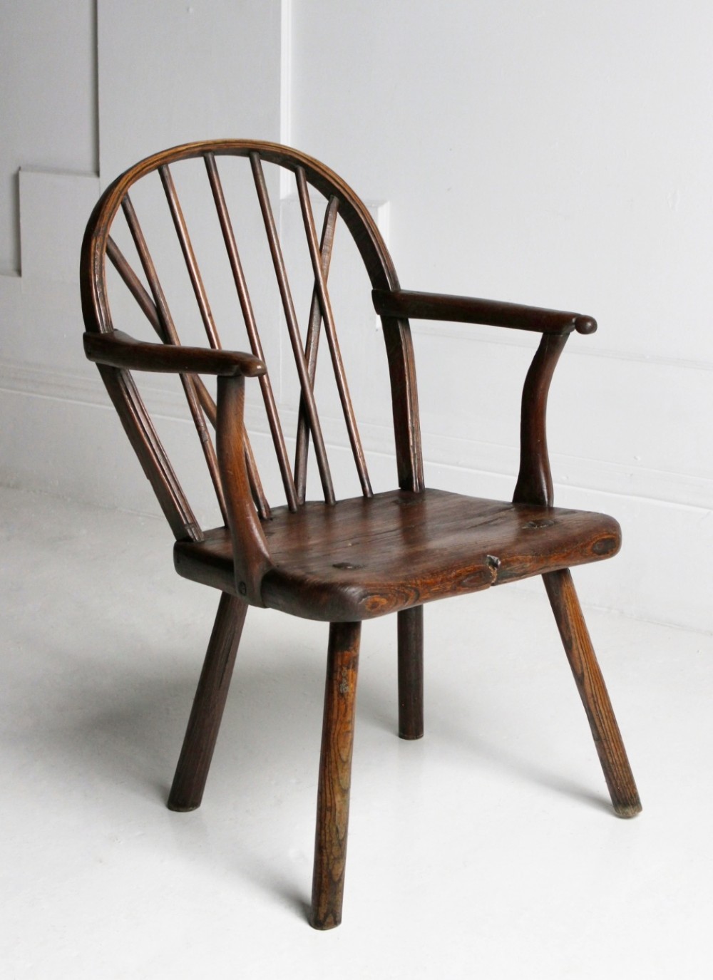 An 18th-century vernacular dark wooden armchair featuring a stick back design, curved armrests, a solid slab seat, and splayed legs.