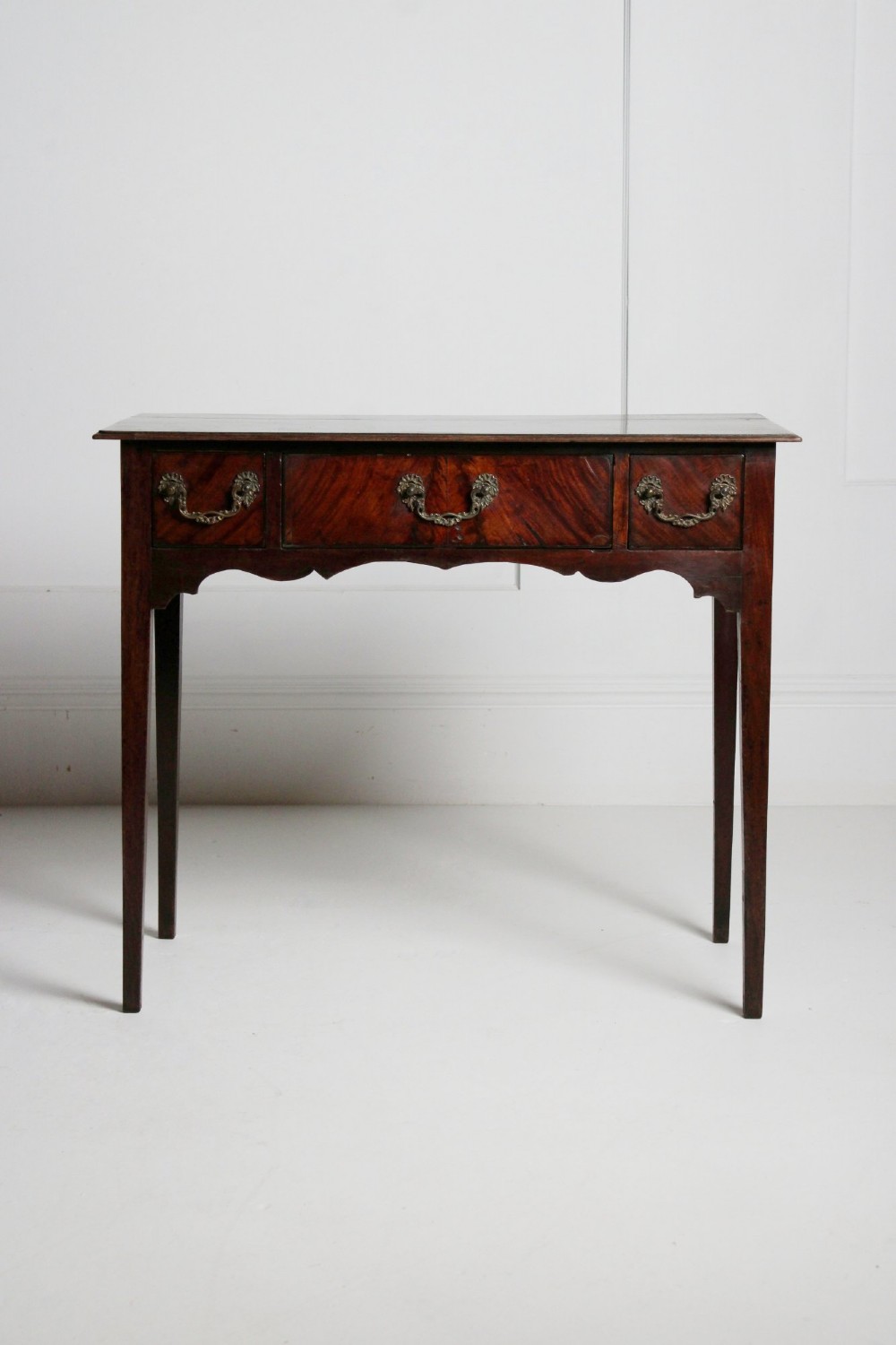 An 18th-century Georgian mahogany side table with three frieze drawers, brass swan-neck handles, a shaped apron, and tapered square legs.