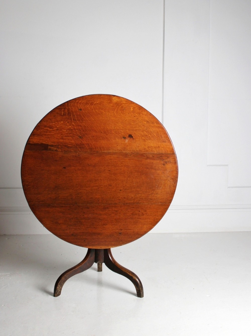 An 18th-century circular oak tilt-top table shown in a vertically tilted position on a turned tripod base.