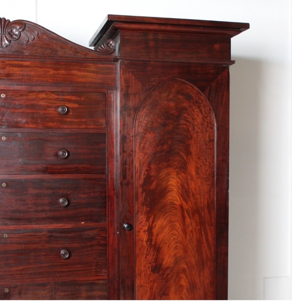 A nineteenth-century Victorian flame mahogany breakfront compactum wardrobe featuring a central chest of drawers flanked by two arched-panel cupboard doors beneath a carved central cresting. View II