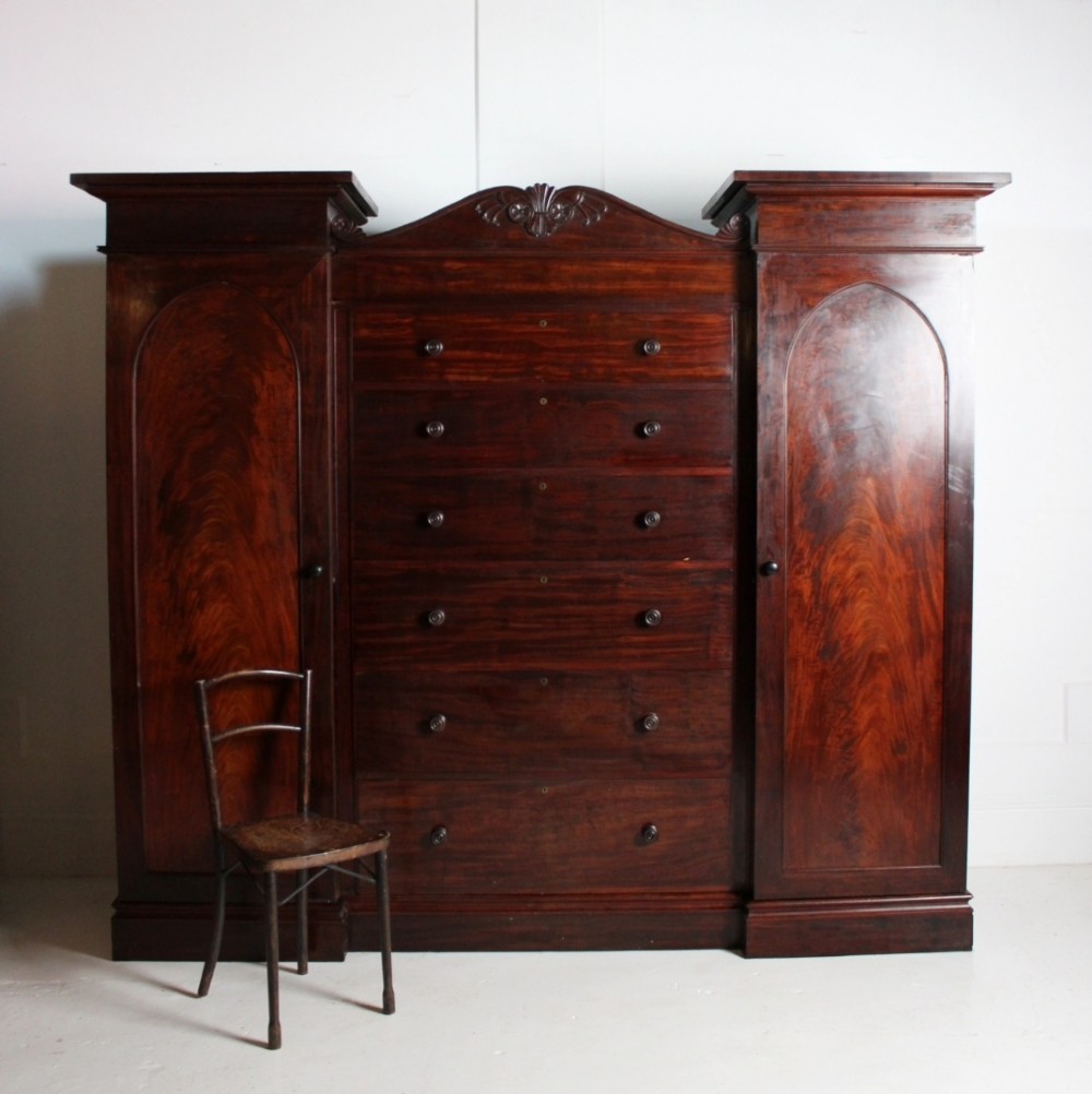 A nineteenth-century Victorian flame mahogany breakfront compactum wardrobe featuring a central chest of drawers flanked by two arched-panel cupboard doors beneath a carved central cresting.