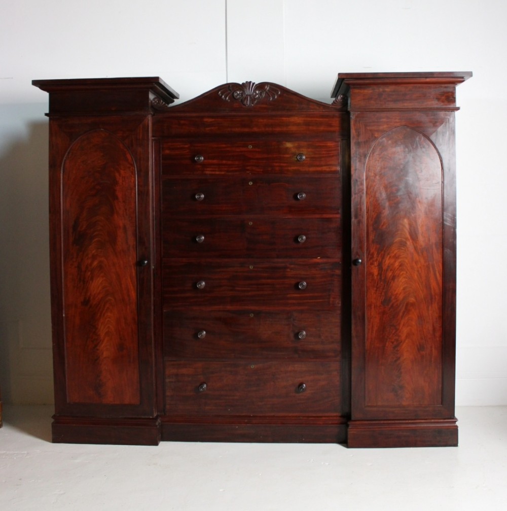 A nineteenth-century Victorian flame mahogany breakfront compactum wardrobe featuring a central chest of drawers flanked by two arched-panel cupboard doors beneath a carved central cresting. View V