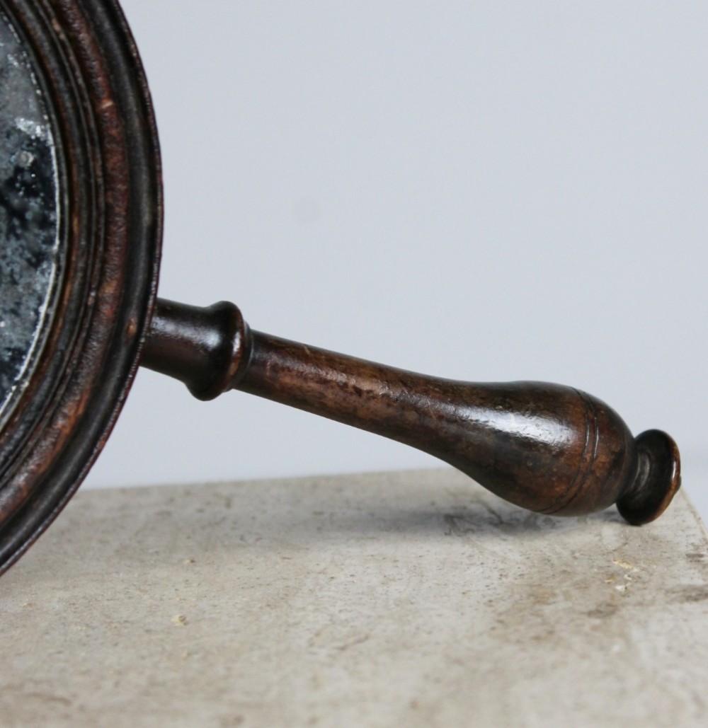 A circular Georgian wooden hand mirror with a turned handle, dark brown finish, top brass hanging loop, and foxed mirror glass. View VI