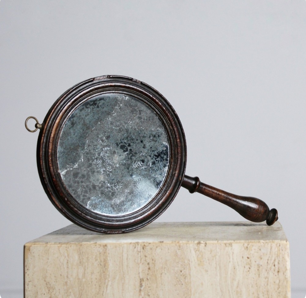 A circular Georgian wooden hand mirror with a turned handle, dark brown finish, top brass hanging loop, and foxed mirror glass.