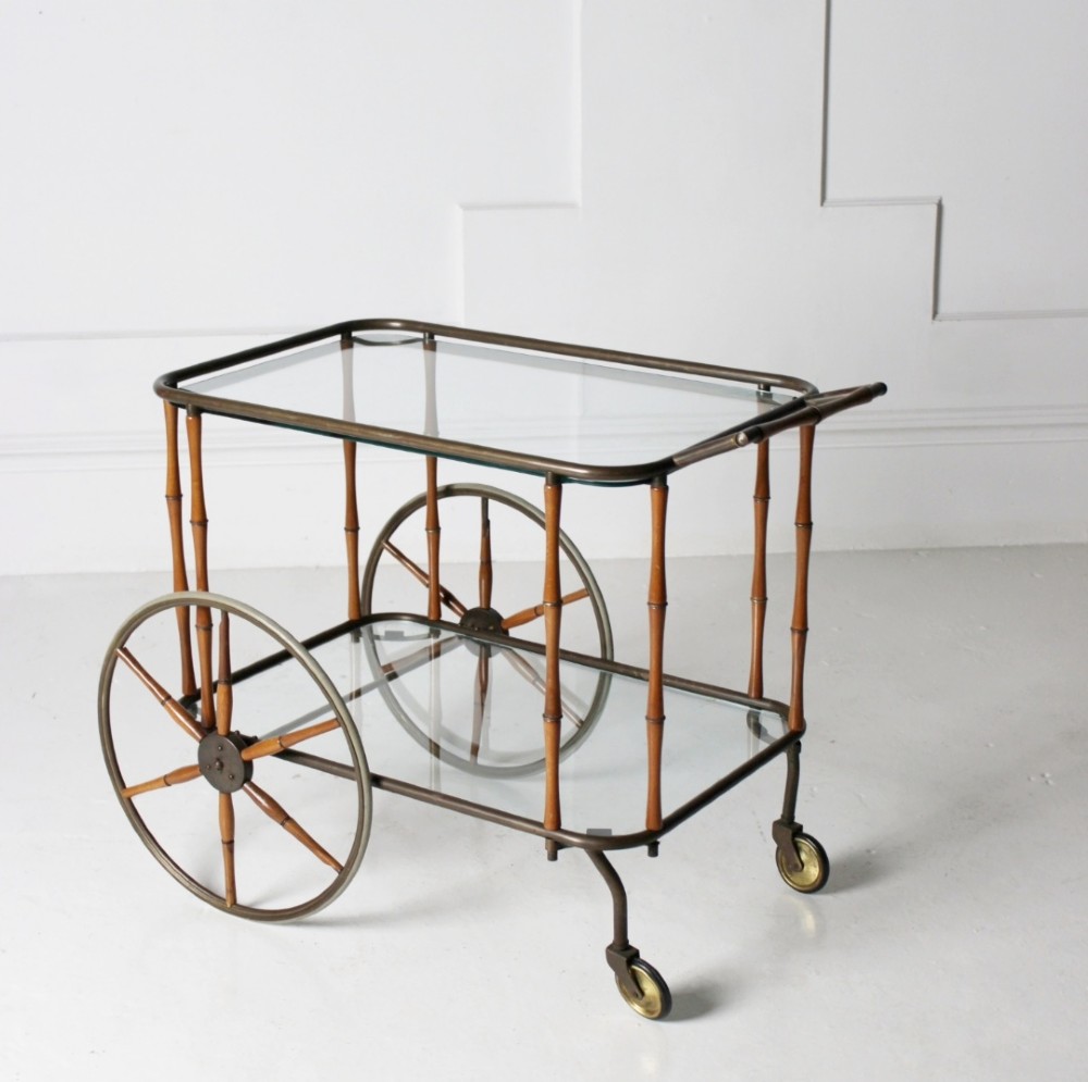 A two-tier mid-century drinks trolley featuring a faux bamboo brass and wood frame, clear glass shelves, two large spoked wheels, and small rear castors.