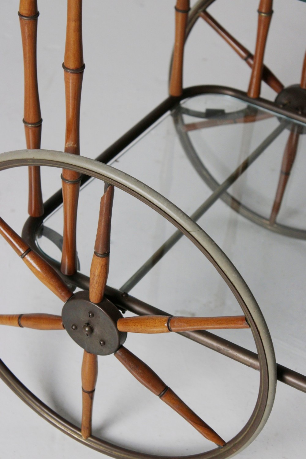 A two-tier mid-century drinks trolley featuring a faux bamboo brass and wood frame, clear glass shelves, two large spoked wheels, and small rear castors. View III