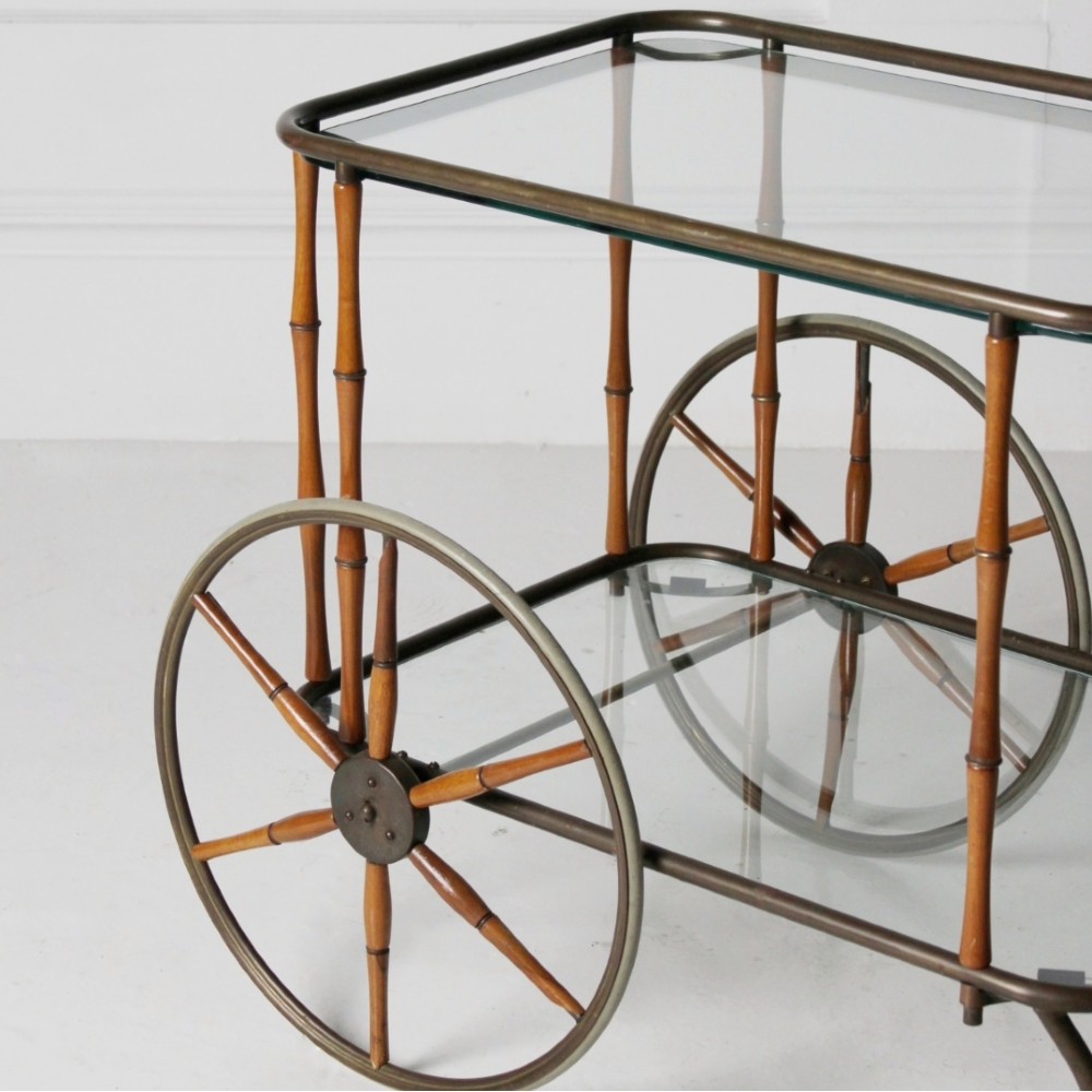 A two-tier mid-century drinks trolley featuring a faux bamboo brass and wood frame, clear glass shelves, two large spoked wheels, and small rear castors. View VI