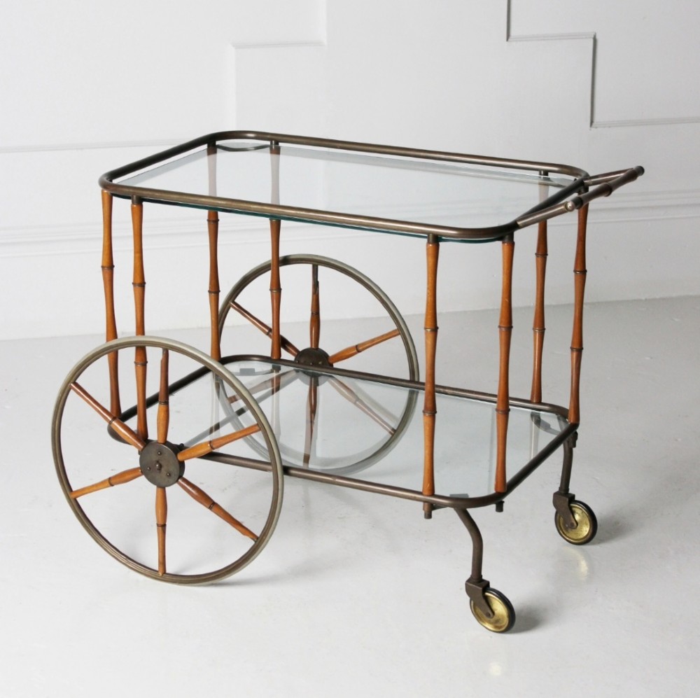 A two-tier mid-century drinks trolley featuring a faux bamboo brass and wood frame, clear glass shelves, two large spoked wheels, and small rear castors. View VII