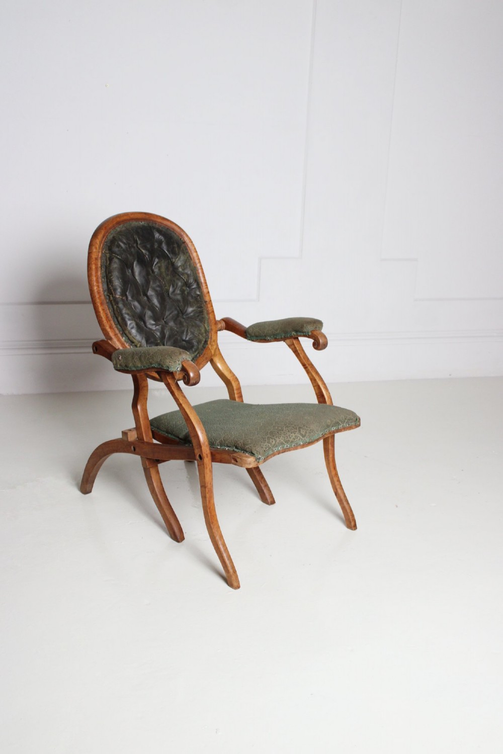 An antique birds eye maple folding armchair featuring curved legs, padded armrests, an oval backrest, and green textured upholstery.