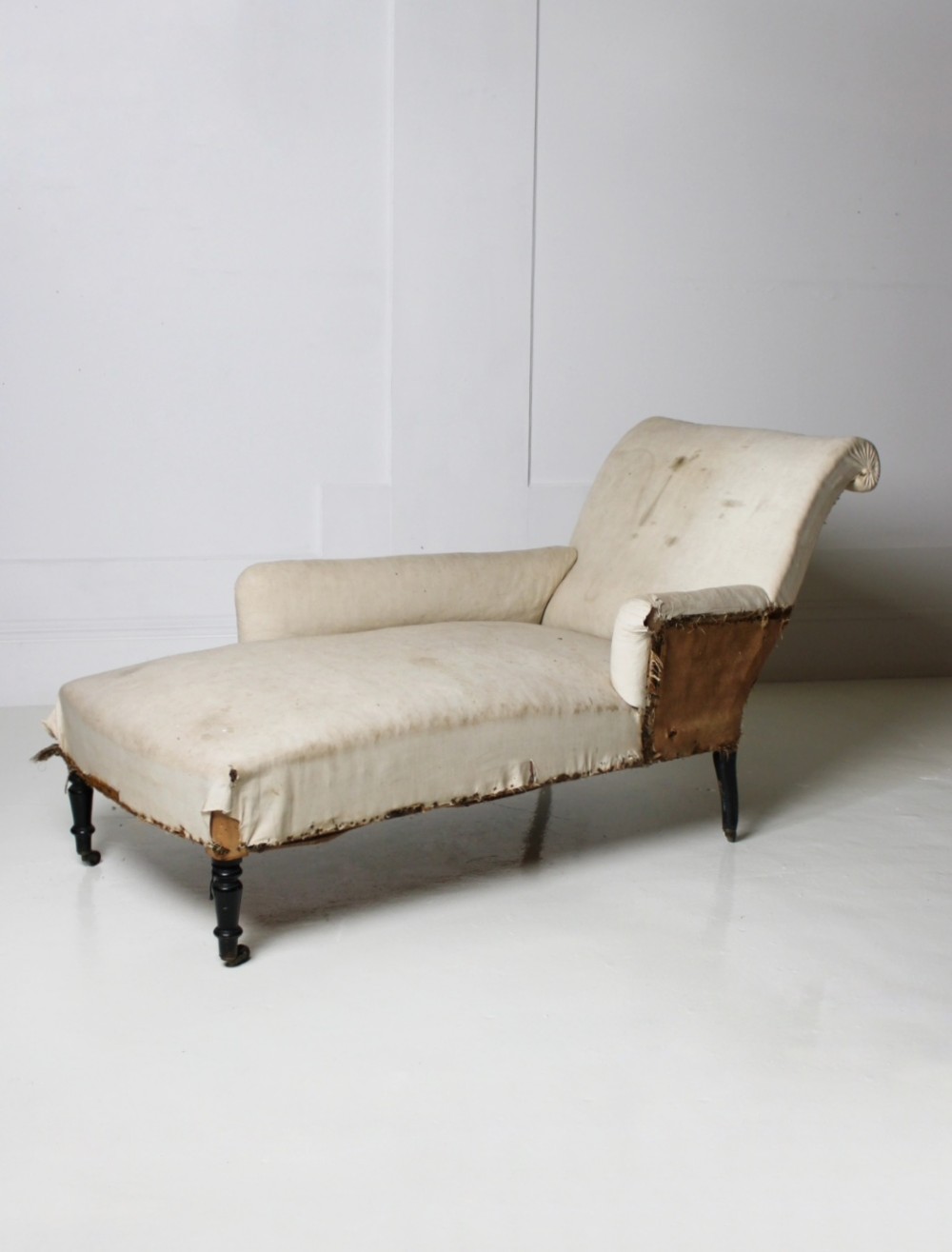 A Victorian country house daybed upholstered in cream calico, featuring turned ebonised legs on brass castors and a scrolled backrest.