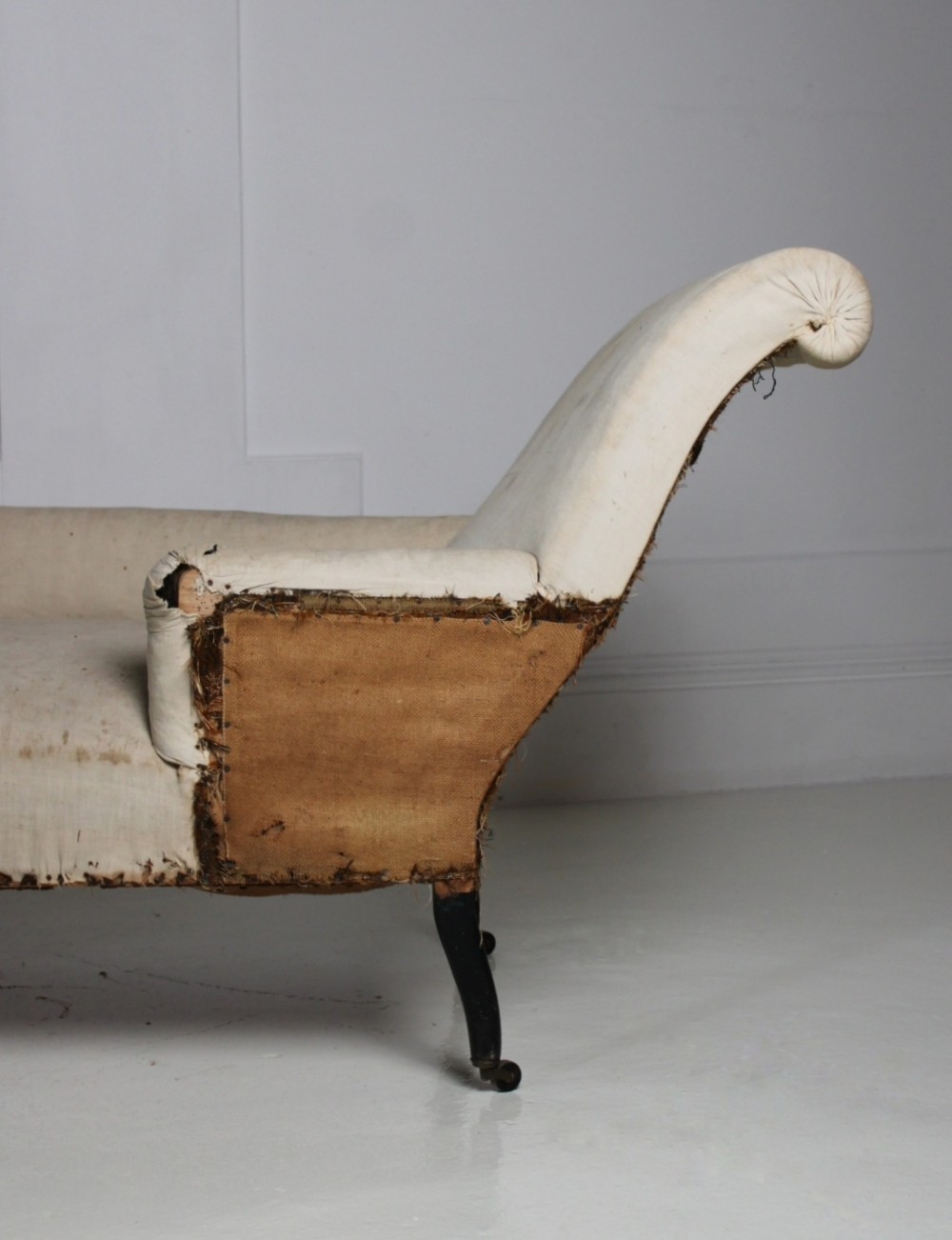 A Victorian country house daybed upholstered in cream calico, featuring turned ebonised legs on brass castors and a scrolled backrest. View IV