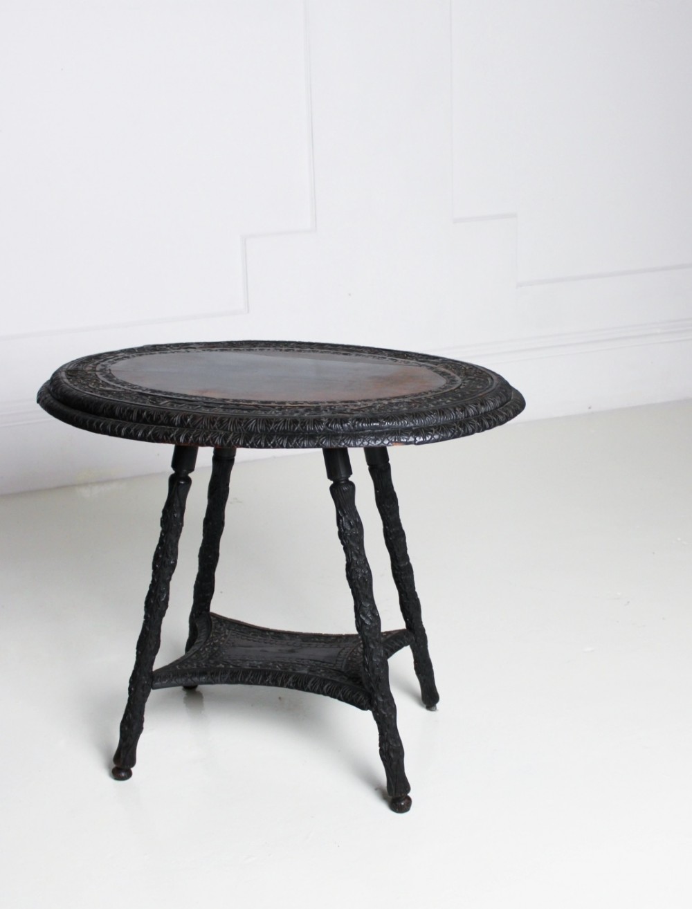 An oval dark timber Anglo-Indian side table featuring hand-carved floral borders, four textured legs, and a carved lower undertier shelf.