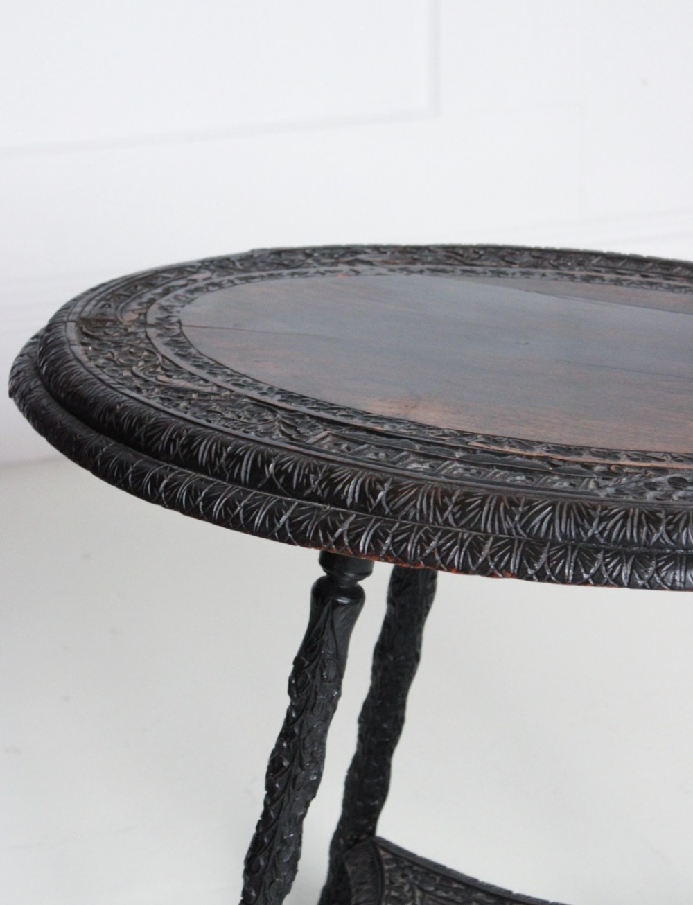 An oval dark timber Anglo-Indian side table featuring hand-carved floral borders, four textured legs, and a carved lower undertier shelf. View III