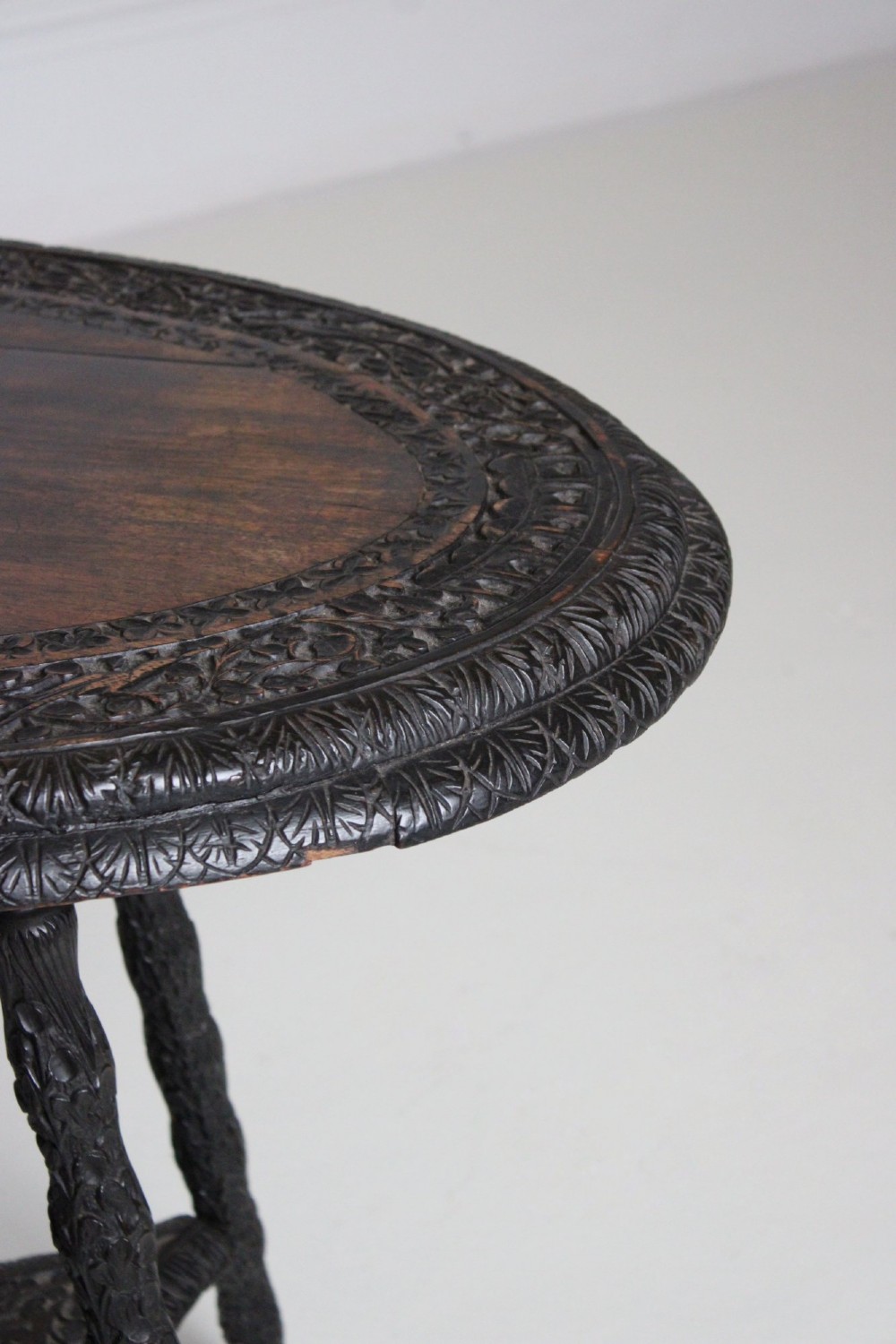 An oval dark timber Anglo-Indian side table featuring hand-carved floral borders, four textured legs, and a carved lower undertier shelf. View VI