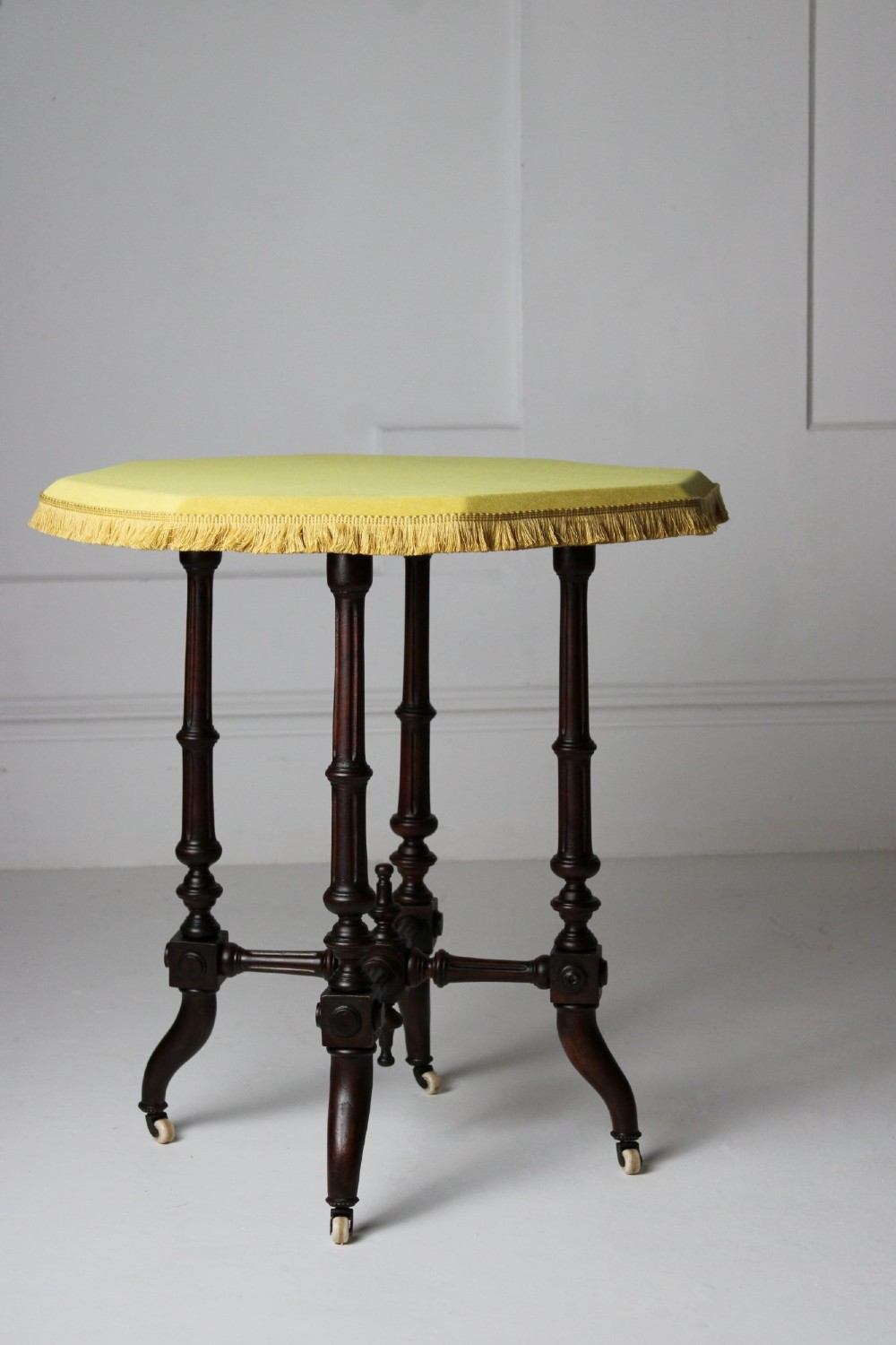 A dark timber Aesthetic Movement side table featuring turned legs, splayed feet on castors, an intricate lower stretcher, and a yellow fabric-covered top finished with fringe trim.