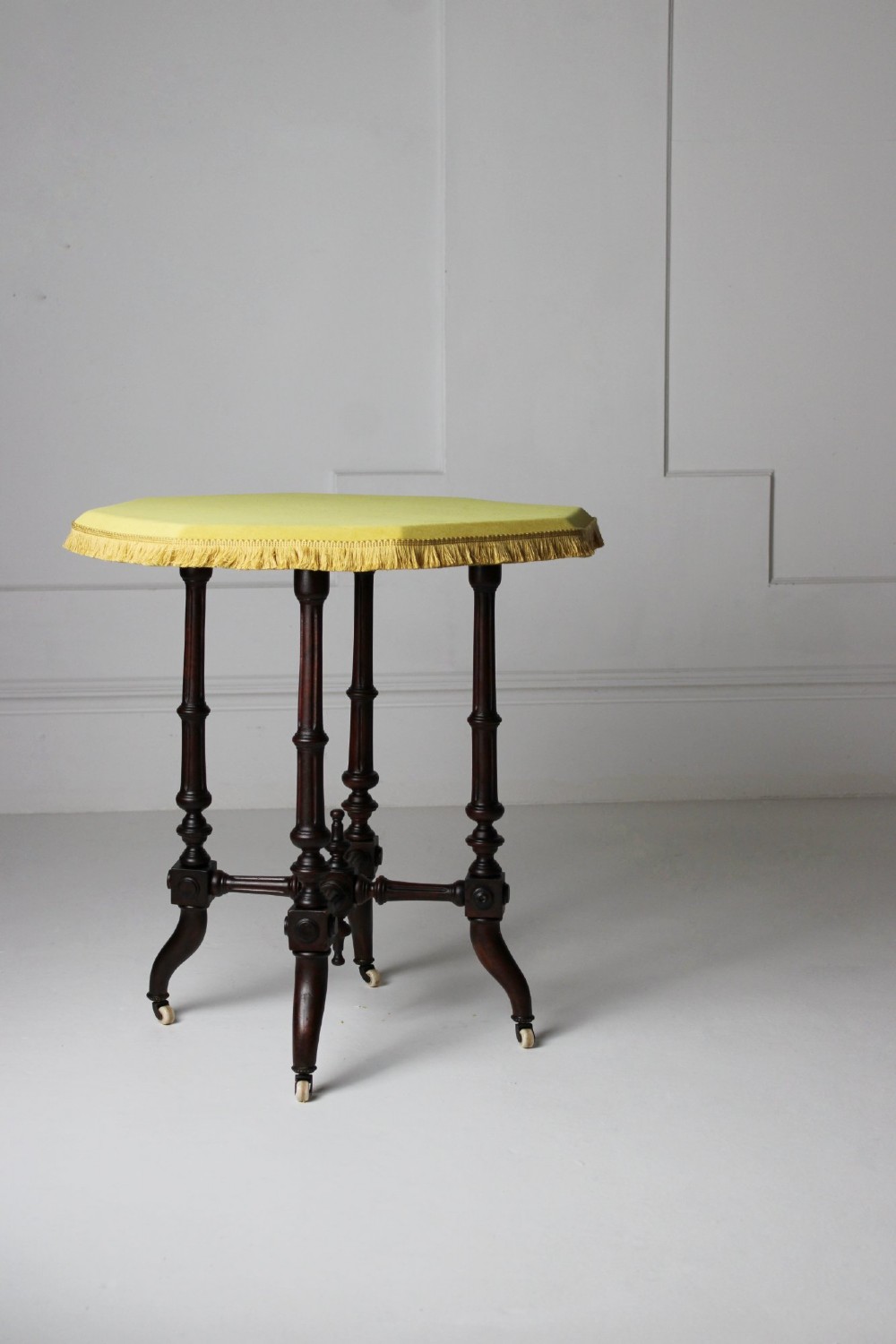 A dark timber Aesthetic Movement side table featuring turned legs, splayed feet on castors, an intricate lower stretcher, and a yellow fabric-covered top finished with fringe trim. View II