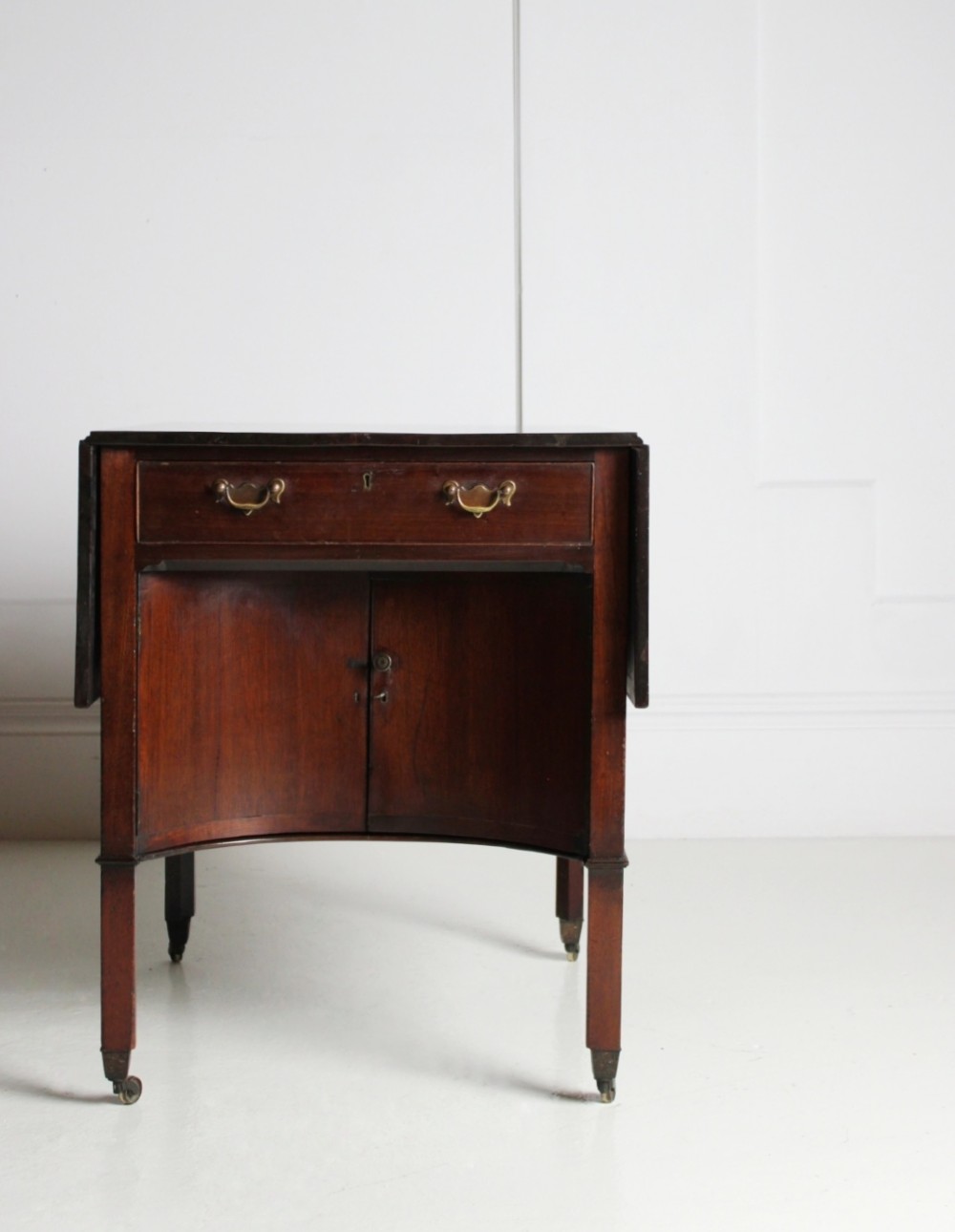 A George III mahogany side table featuring drop leaves, a single top drawer with brass handles, a concave lower cupboard, and square tapered legs on castors.
