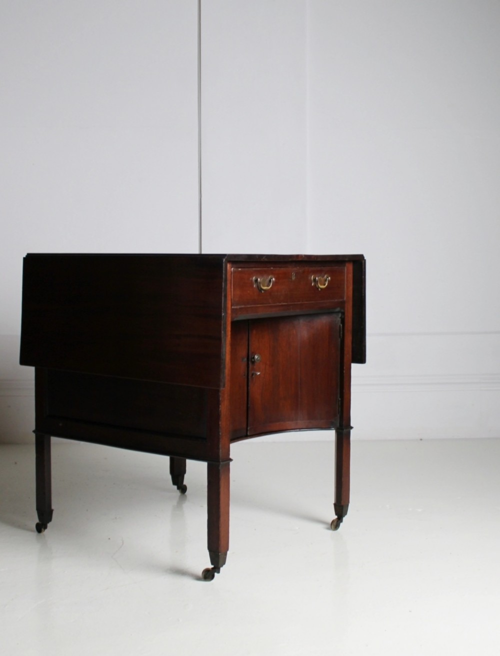 A George III mahogany side table featuring drop leaves, a single top drawer with brass handles, a concave lower cupboard, and square tapered legs on castors. View II