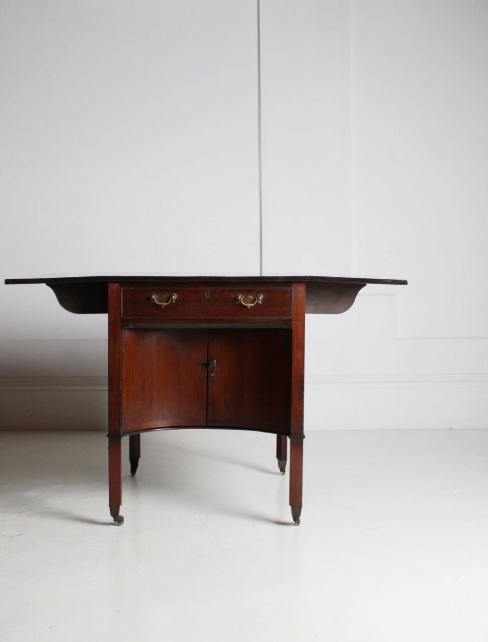 A George III mahogany side table featuring drop leaves, a single top drawer with brass handles, a concave lower cupboard, and square tapered legs on castors. View V