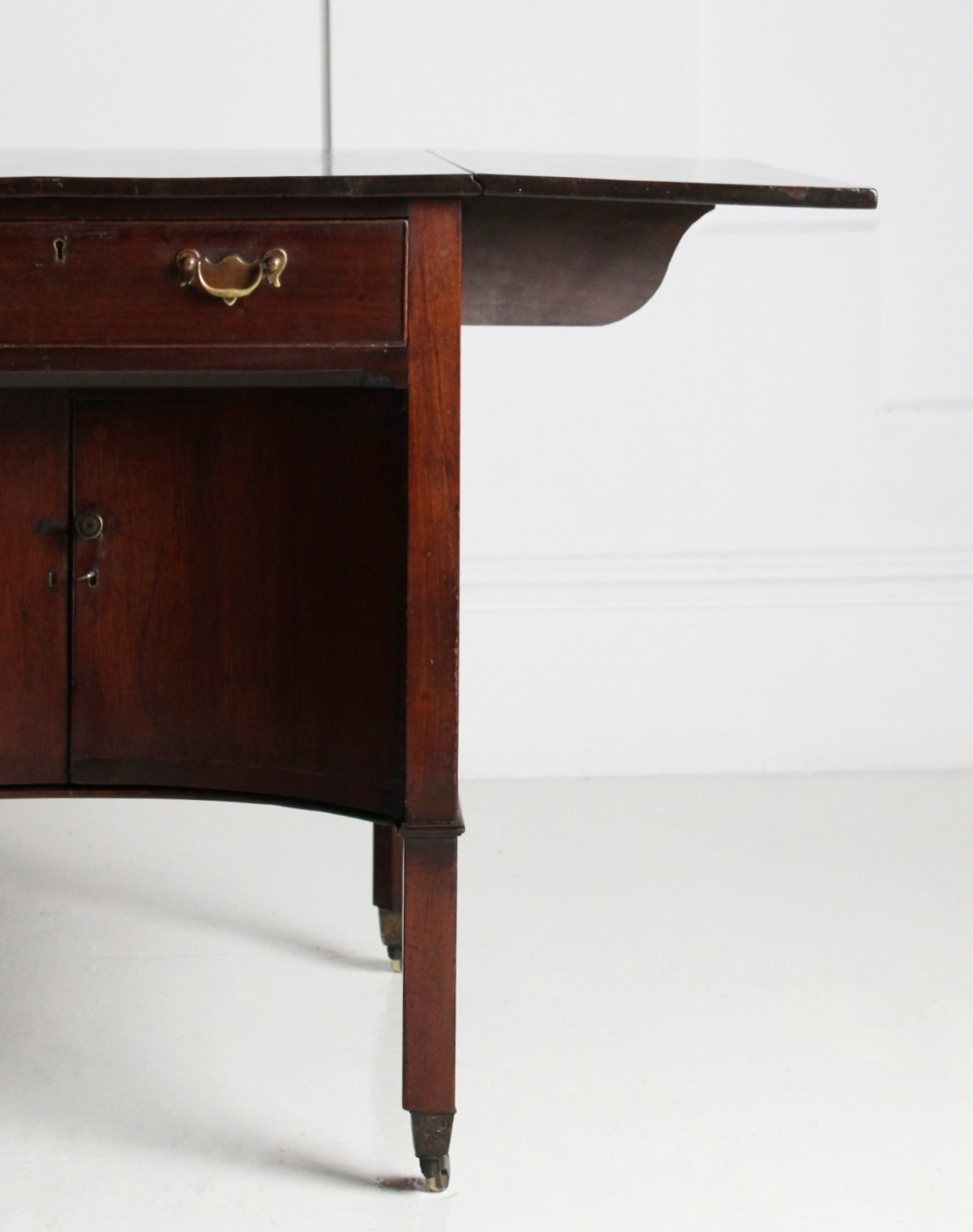 A George III mahogany side table featuring drop leaves, a single top drawer with brass handles, a concave lower cupboard, and square tapered legs on castors. View VI