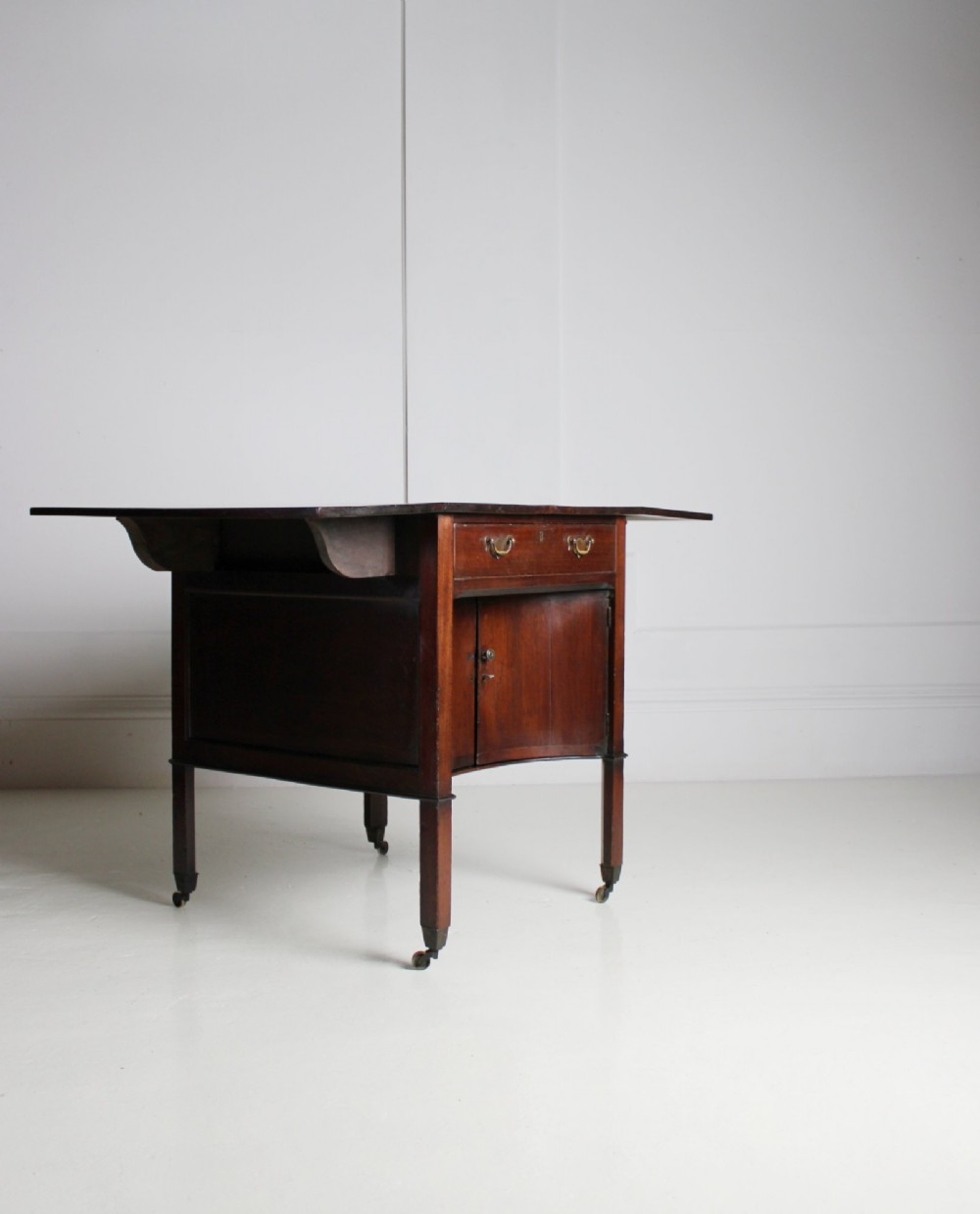A George III mahogany side table featuring drop leaves, a single top drawer with brass handles, a concave lower cupboard, and square tapered legs on castors. View VII