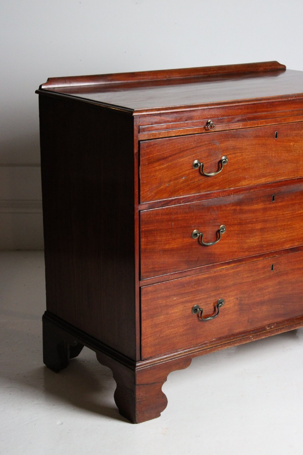 A George III mahogany chest of drawers featuring three graduated long drawers with brass swan-neck handles and bracket feet. View II