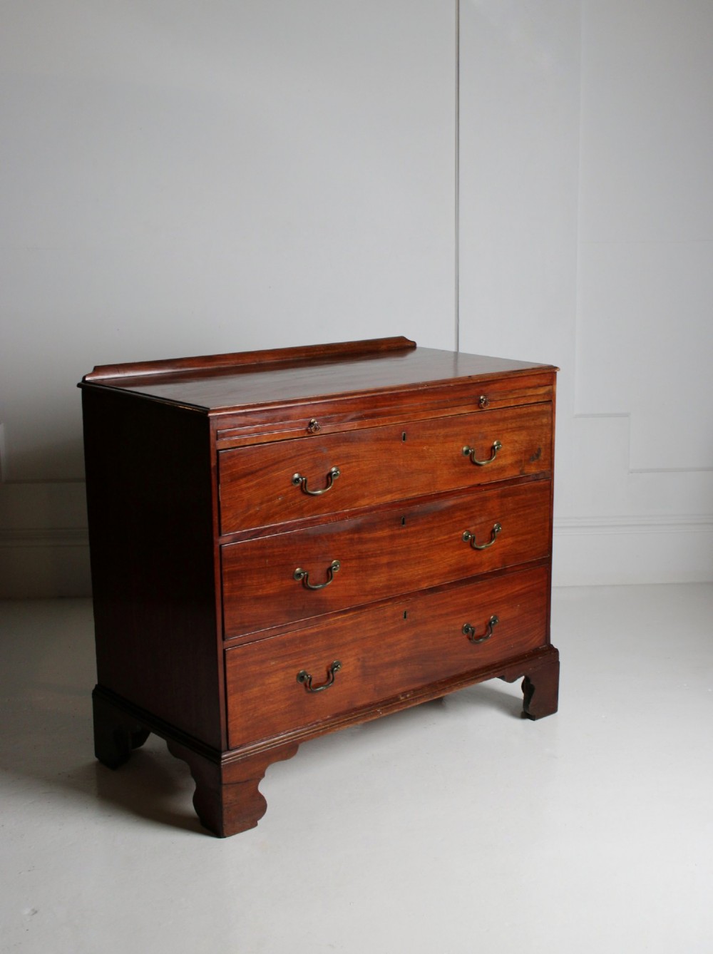 A George III mahogany chest of drawers featuring three graduated long drawers with brass swan-neck handles and bracket feet. View IV
