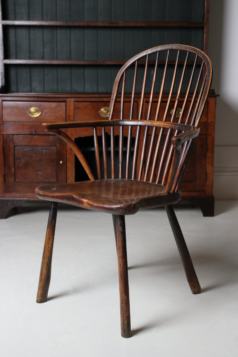 An 18th-century vernacular wooden Windsor armchair featuring a steam-bent hoop back, slender spindles, a carved saddle seat, and splayed legs.