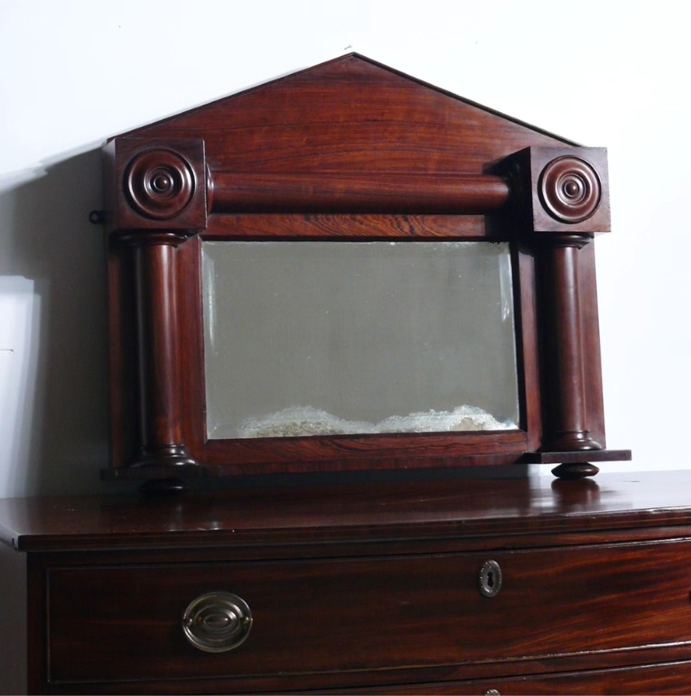 An antique Regency rosewood overmantel mirror featuring a triangular pediment, carved circular roundels, and flanking turned columns surrounding a rectangular mirror plate.