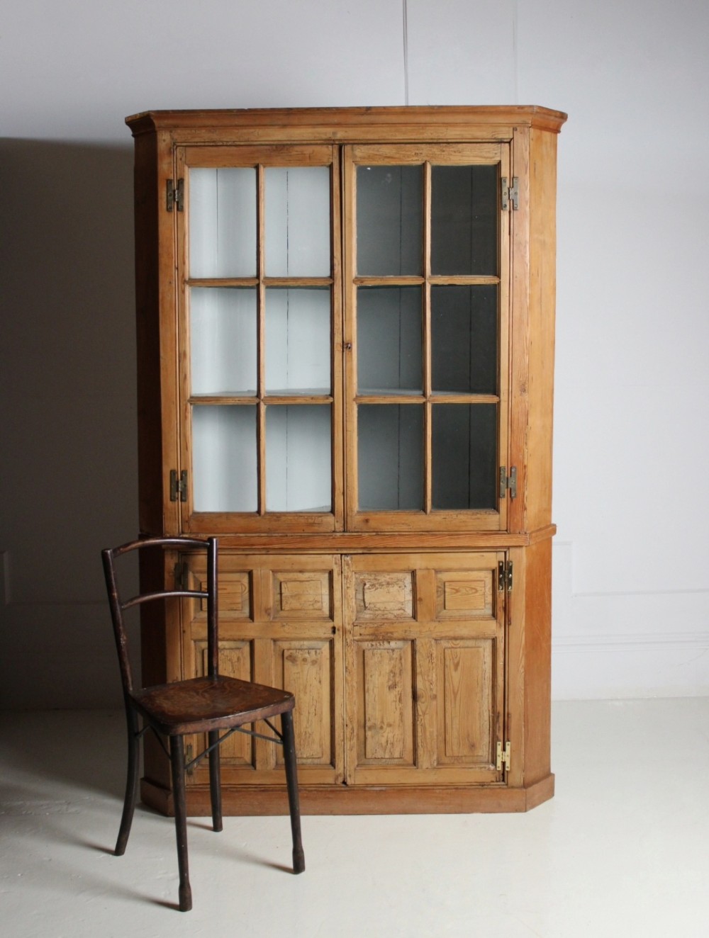 A tall freestanding pine corner cupboard featuring a glazed upper display section with multi-pane glass doors above a two-door lower cabinet with raised timber panels.