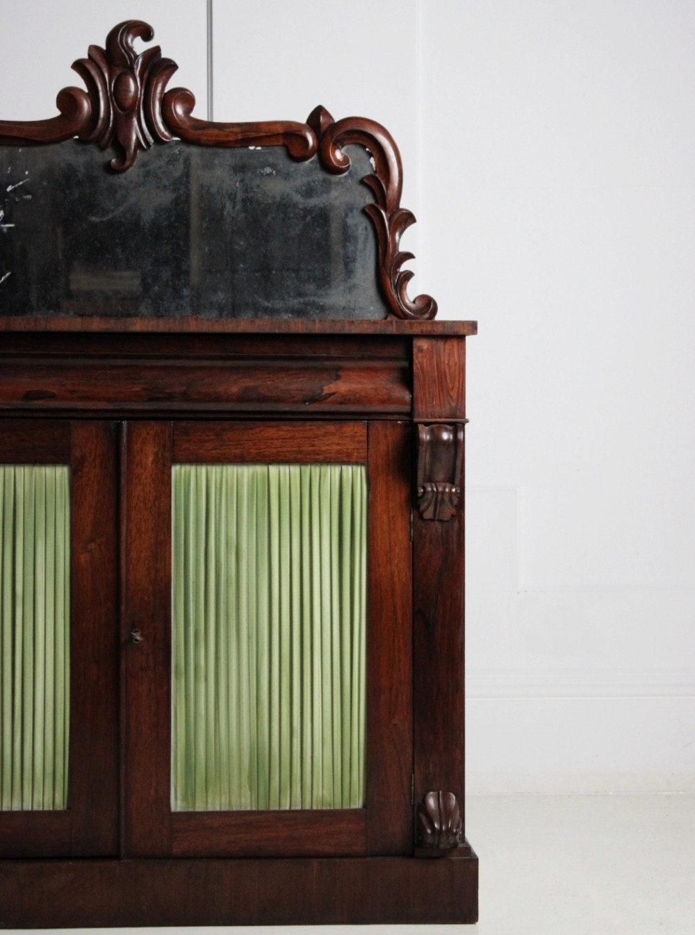 A Regency rosewood chiffonier featuring a carved crest above a distressed mirror back and two lower doors with pleated green fabric panels. View II