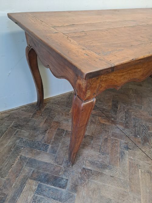 fig 1. An early 19th-century French farmhouse kitchen table made of warm brown wood with a plank top, scalloped apron, and curved cabriole legs.