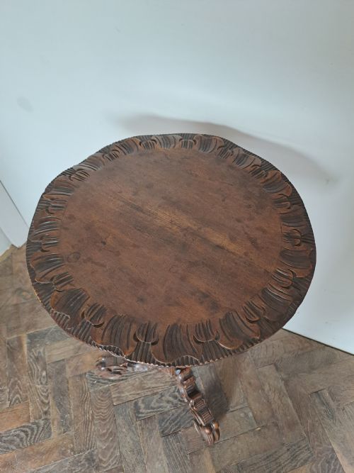 fig 1. A dark brown carved wood tripod table featuring an ornate scalloped rim top, a detailed central pedestal, and three scrolled legs.