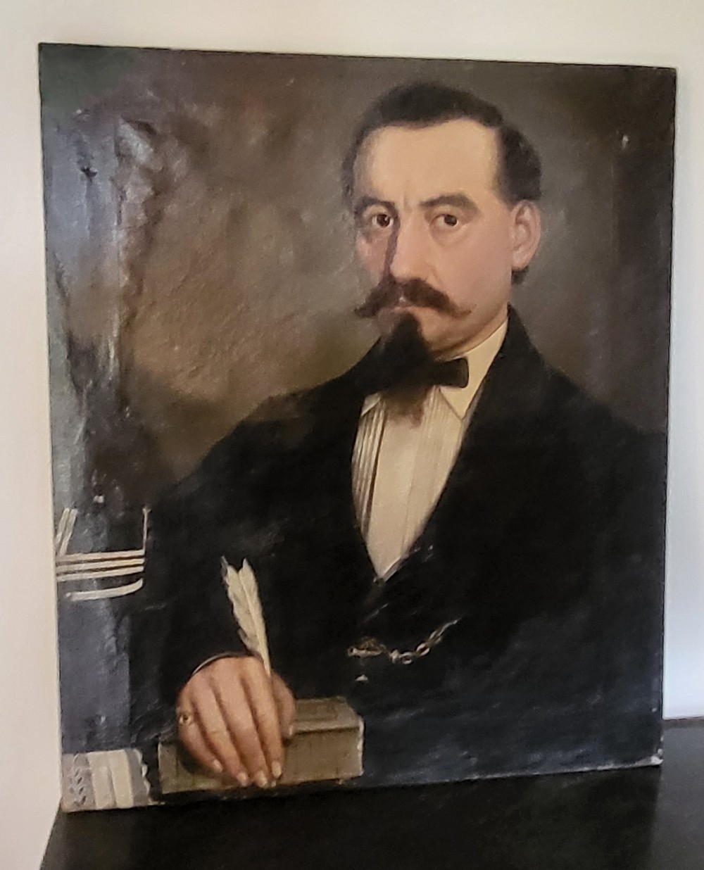 An antique oil painting on canvas depicting a gentleman with a moustache holding a feather quill pen over a book.