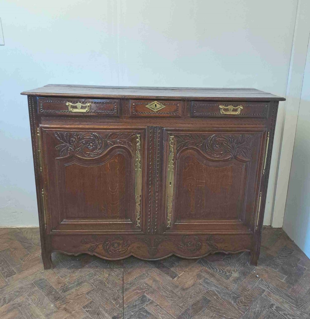 A carved dark oak French provincial buffet base featuring two relief-carved cupboard doors with long brass hinges below two drawers.