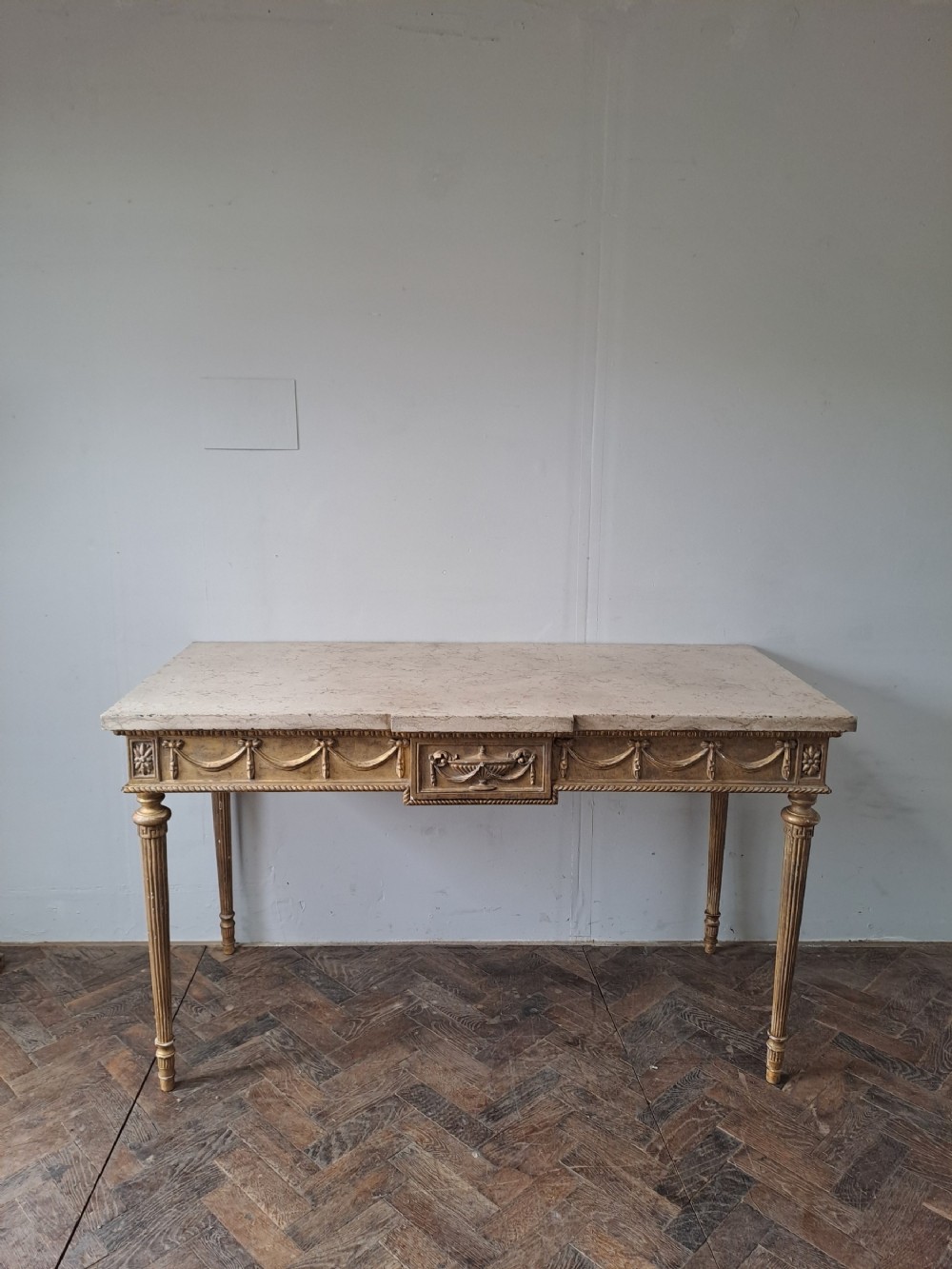 A French Louis XVI style giltwood console table with a cream marble top, featuring carved swags, an urn motif, and four fluted legs.