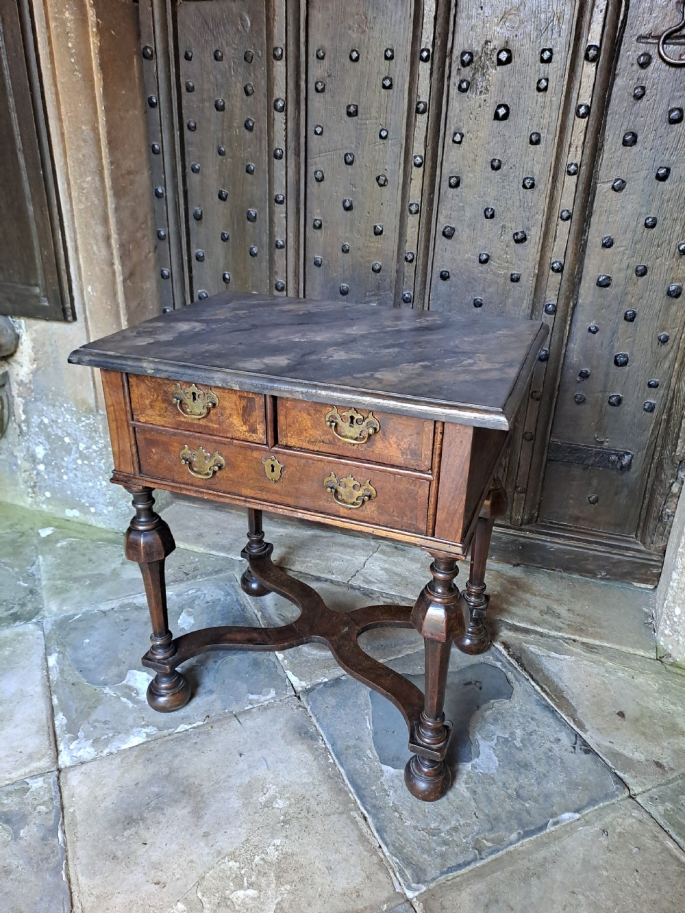 A walnut side table featuring three drawers with brass handles, inverted cup turned legs, and a curved X-stretcher.